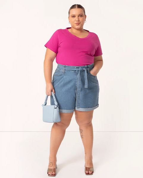 modelo usa short plus size clochard jeans cintura super alta com faixa azul escuro, blusa rosa e bolsa