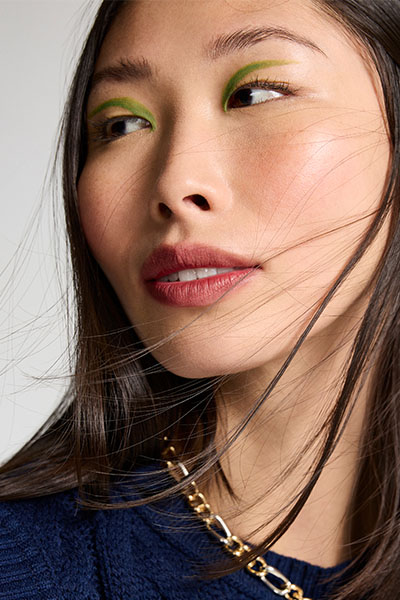 Modelo asiática usa delineador gráfico verde e veste blusa de tricô azul marinho e corrente dourada no pescoço