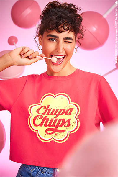 Modelo de cabelos cacheados pisca um olho só e sorri, com pirulito na boca e vestindo uma camiseta vermelha com o logo de Chupa Chups