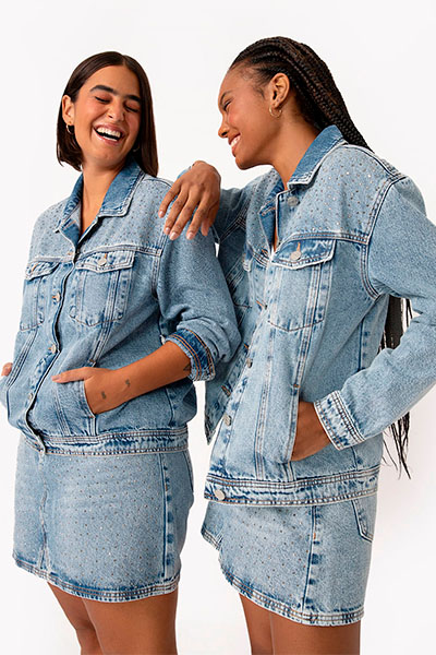 Sorrindo e apoiadas uma na outra, modelos vestem o mesmo look: jaqueta jeans e saia jeans, ambas as peças têm aplicação de strass nas suas partes superiores