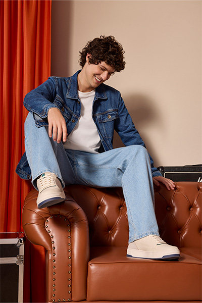 Modelo está sentado no braço e encosto de um sofá de couro caramelo. Ele veste jaqueta jeans em lavagem regular e calça wide leg em lavagem mais clara com tênis branco. 