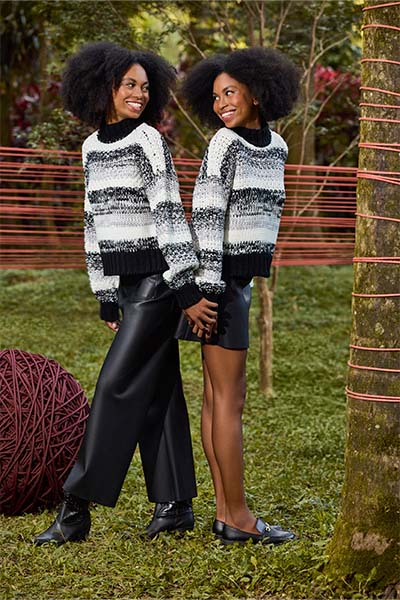 Modelos vestem o mesmo suéter de tricô listrado preto e branco e texturizado. a Modelo da esquerda está vestindo calça preta de PU e bota preta, enquanto a segunda veste saia de PU preta e mocassim. Elas estão em um parque