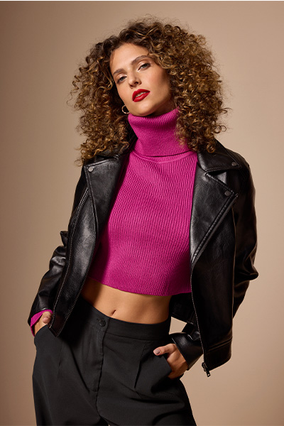 Modelo veste blusa cropped de tricô pink com gola alta, jaqueta de PU por cima e calça parachute preta.