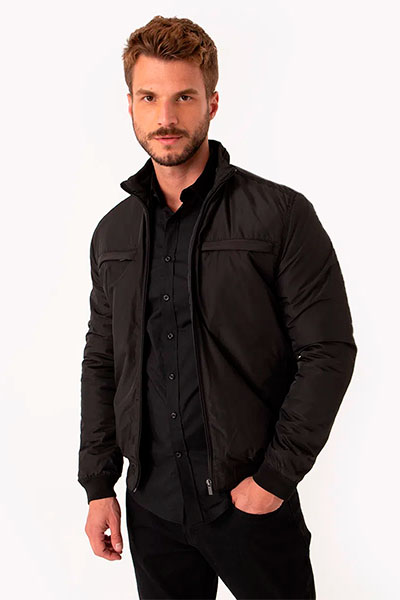 Modelo veste jaqueta de nylon preta com camiseta polo preta por baixo e calça preta.