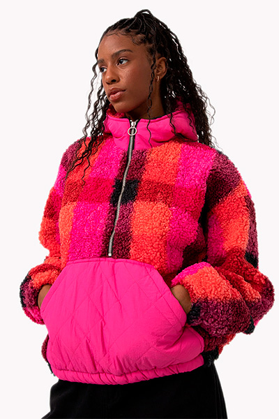 Modelo veste casaco sherpa xadrez, em tons de pink, preto e laranja, com bolso canguru todo em pink, e calça preta