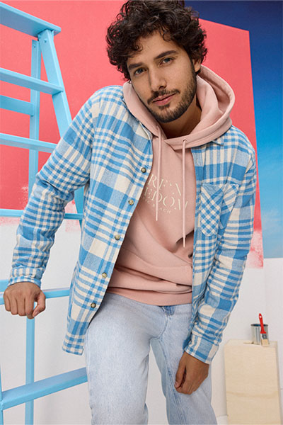 Modelo veste jaqueta xadrez azul e branca por cima de moletom rosa pastel e calça jeans de lavagem clara