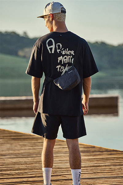 De costas, modelo veste boné branco, camiseta preta com o escrito "a fisherman's tale" nas costas, bermuda preta e tênis branco