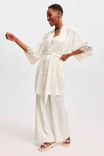 Modelo veste kimono branco, calça pantalona branca e sandália branca. As peças foram feitas em parceria com a marca Demodê e fazem parte da coleção Nosso Encontro 2023