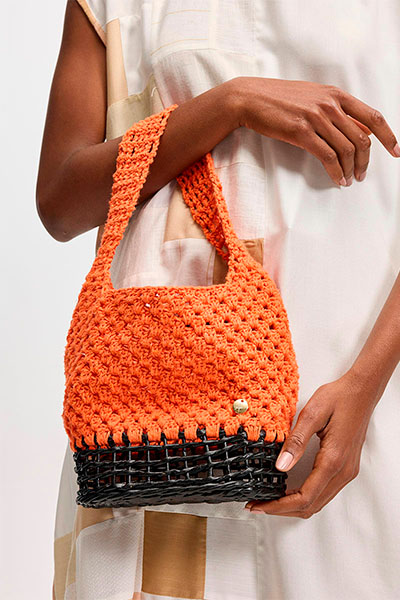 Modelo segura no braço bolsa de crochê laranja com a parte de baixo preta. Peça foi feita em parceria com a marca Casa de Maria e faz parte da coleção Nosso Encontro 2023