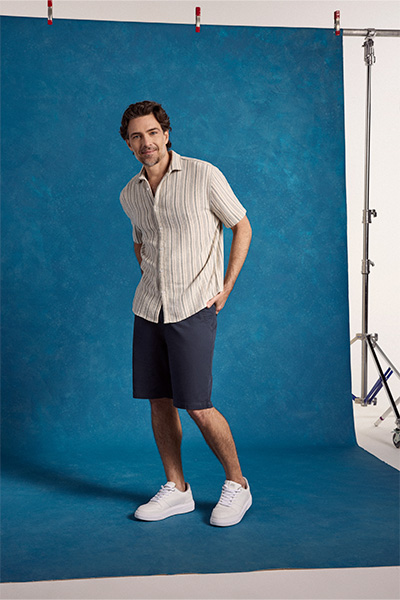 Modelo veste camisa de manga curta de algodão com listras verticais, branco e off white, bermuda chino azul marinho e tênis branco