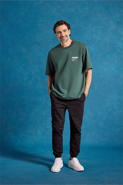 Modelo veste camiseta verde escura, calça azul marinho e tênis branco