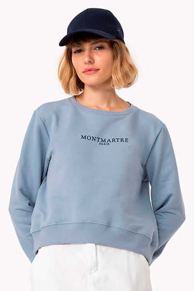 Modelo veste moletom azul claro com o escrito "Montmartre Paris", calça branca e boné azul marinho