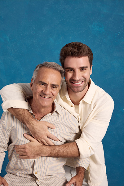 O influenciador digital Diego Gondim está abraçando seu pai, Danilo. Diego veste camisa de manga longa off white e bermuda chino off white. Danilo, o pai, veste camisa manga longa cru