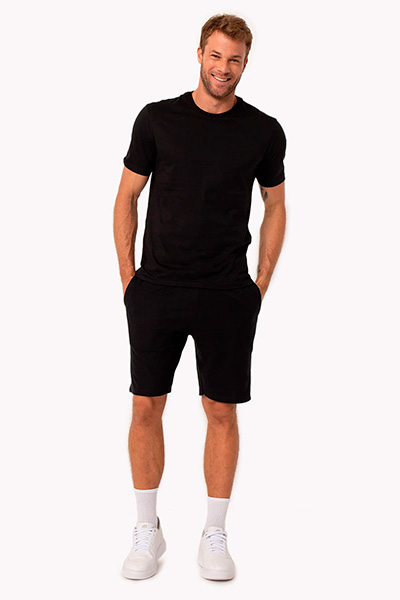 Modelo veste camiseta preta, bermuda preta, meia branca e tênis branco
