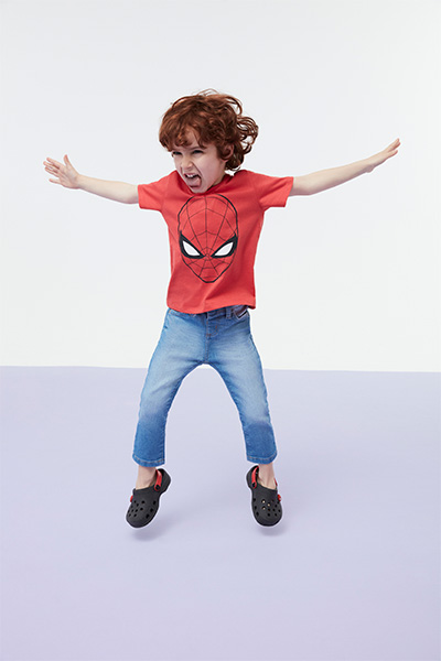 Menino veste camiseta infantil vermelha do Homem Aranha e calça jeans com tênis preto