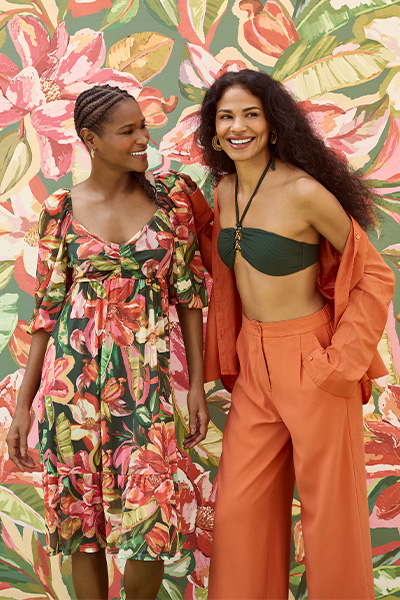 Da esquerda para a direita, modelos vestem vestido estampado com fundo verde escuro e flores em rosa, vermelho e amarelo; e top verde com chemise laranja aberta por cima e calca de alfaiataria laranja no mesmo tom