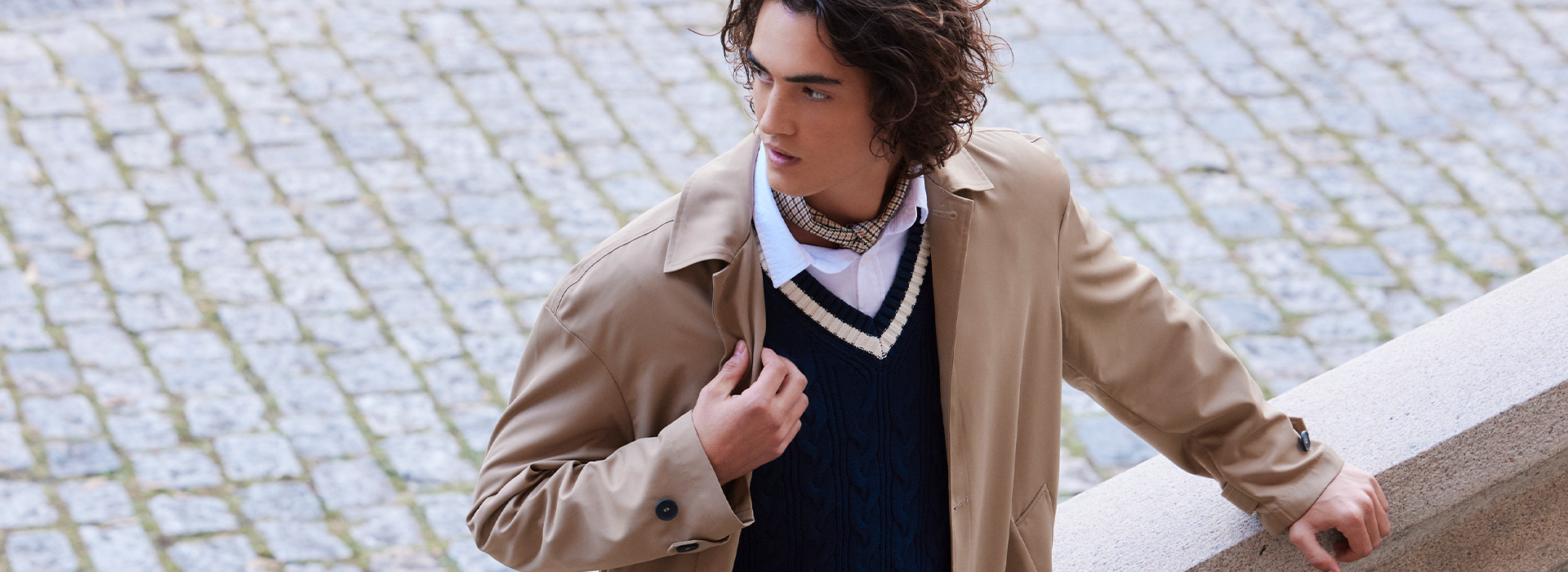 Modelo usando um look de inverno com camadas: camisa social, colete de tricô e trench coat por cima.
