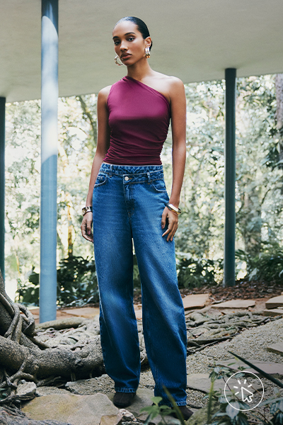 Modelo veste blusa assimétrica de um ombro só vinho, máxi brincos de argola dourados e calça jeans baggy azul médio
