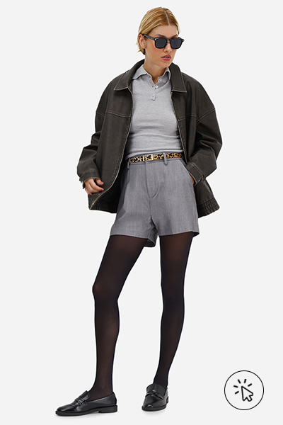 Modelo veste jaqueta jeans oversized preta, polo cinza, short de alfaiataria feminino com cinto cinza, meia-calça preta e mocassim