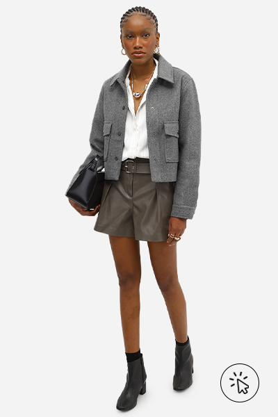 Modelo veste jaqueta estruturada cinza por cima de camisa branca feminina, short feminino de couro PU com cinto cinza e botas de cano curto e salto bloco pretas