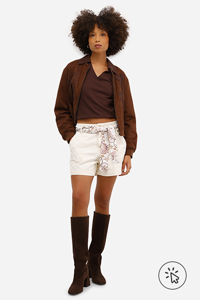 Modelo veste jaqueta de suede marrom com gola esportiva, blusa gola V cropped marrom, short feminino de sarja com faixa bege e botas over the knee de suede marrom