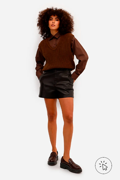 Modelo veste colete feminino de tricô marrom por cima de camisa social na mesma cor, short de PU preto e mocassim preto