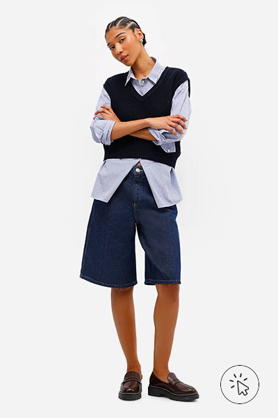 Modelo veste camisa feminina azul clara, colete de tricô azul marinho e bermuda jorts feminina jeans azul