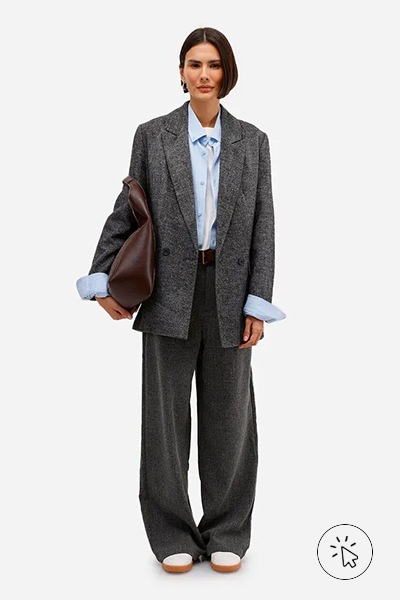 A modelo veste um blazer de alfaiataria cinza oversized combinado com uma camisa social azul clara. A produção é finalizada com calça de alfaiataria wide leg no mesmo tom cinza do blazer, cinto marrom e tênis brancos. Nos acessórios, ela carrega uma maxi bolsa marrom de couro e usa brincos discretos. - roupas anos 80 - look anos 80 - moda anos 80 - como era a moda nos anos 80