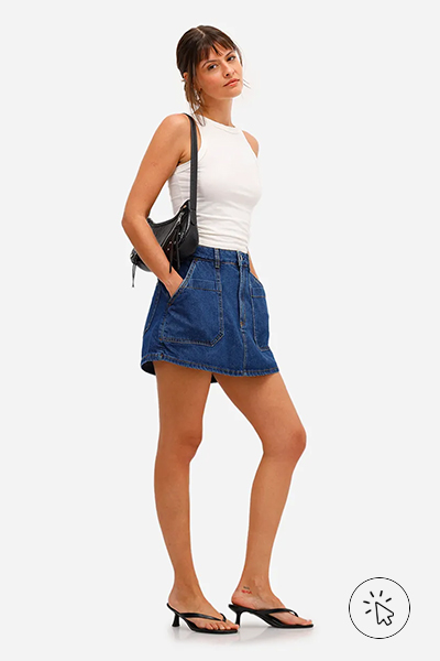 Modelo veste regata off-white justa, minissaia jeans de lavagem escura e modelagem reta, sandálias pretas de salto baixo e bolsa preta tiracolo - história do jeans - como surgiu o jeans - como é feito o tecido jeans - tipos de jeans tecido - modelagens de jeans 