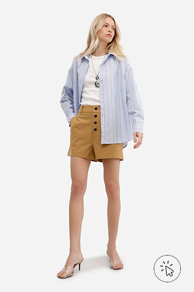 Modelo veste camisa oversized listrada de manga longa off-white, regata off-white, maxi colar prateado, short de alfaiataria caramelo e tamanco de vinil com salto médio.- como usar camisa listrada