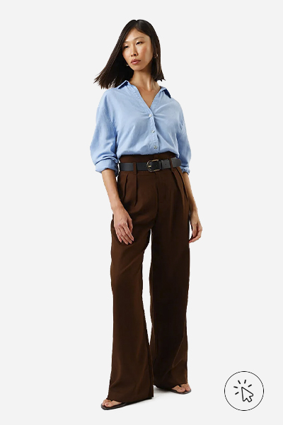 A modelo está usando uma camisa azul, uma calça marrom e um sinto preto.  - tecido linho - o que é linho - como lavar roupa de linho - como passar roupa de linho - como é feito o linho 