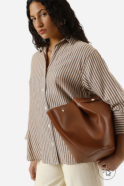 A imagem é só a parte de cima da modelo, ela está usando uma camisa listrada, calça bege e está segurando uma bolsa marrom.  - tecido linho - o que é linho - como lavar roupa de linho - como passar roupa de linho - como é feito o linho 