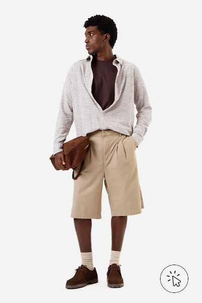 O modelo está usando uma bermuda bege, uma camisa marrom e uma camisa xadrez por cima. Ele tá segurando uma bolsa marrom, meia comprida e mocassim marrom de suede - estilo grunge - o que é o estilo grunge